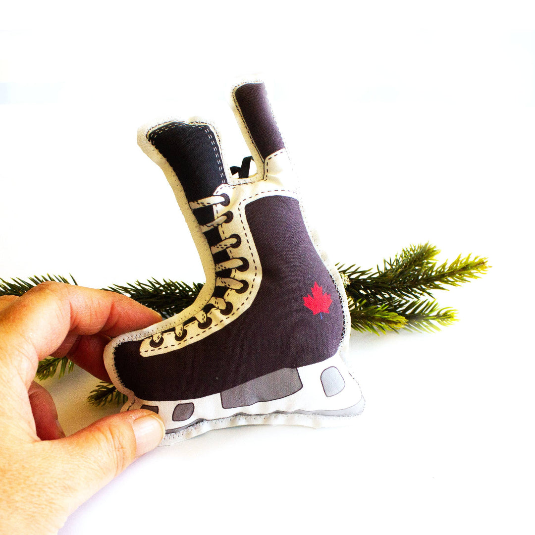 A close-up, high-angle flat lay shot on a plain white background features a soft, plush Christmas ornament shaped like a hockey skate. The skate is black and white, with an illustration of a red maple leaf on the side of the boot. The blade of the skate is grey and white. A red ribbon loop is attached to the top for hanging.