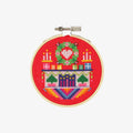 A small, circular embroidery hoop with a festive Christmas scene. The scene depicts a fireplace with candles, presents, and a Christmas tree. The embroidery is colorful and detailed, with a red background.