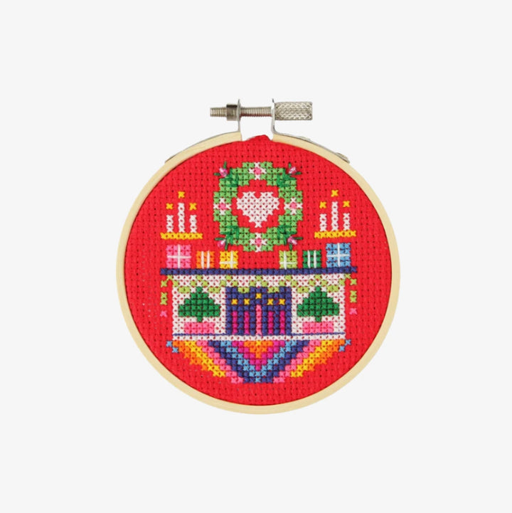 A small, circular embroidery hoop with a festive Christmas scene. The scene depicts a fireplace with candles, presents, and a Christmas tree. The embroidery is colorful and detailed, with a red background.