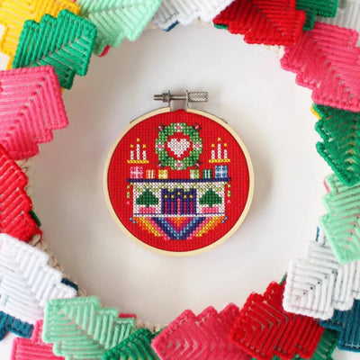 A small, circular embroidery hoop with a festive Christmas scene. The scene depicts a fireplace with candles, presents, and a Christmas tree. The embroidery is colorful and detailed, with a red background.