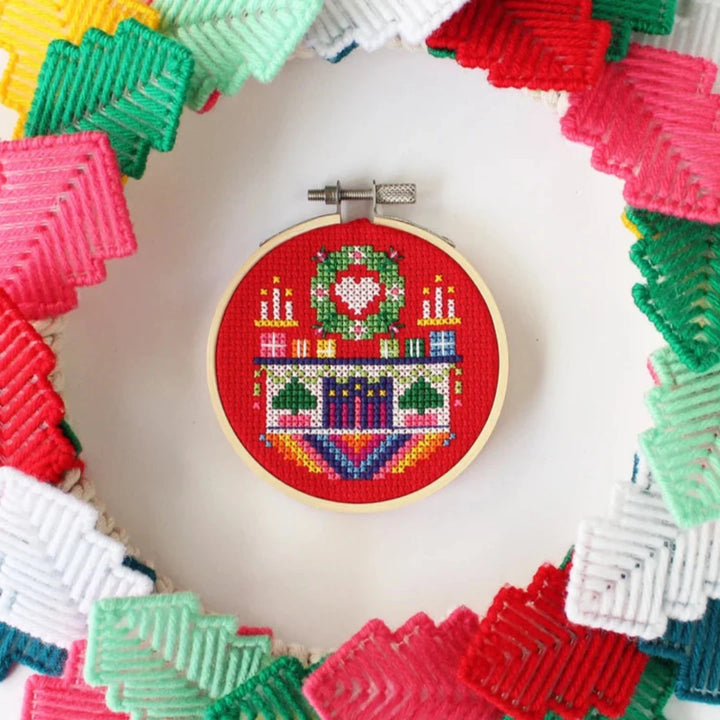 A small, circular embroidery hoop with a festive Christmas scene. The scene depicts a fireplace with candles, presents, and a Christmas tree. The embroidery is colorful and detailed, with a red background.
