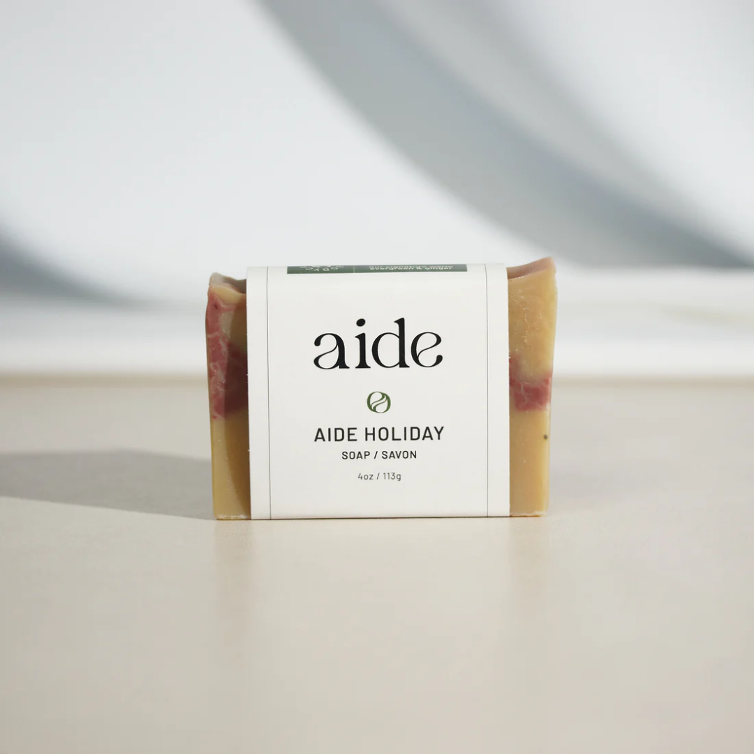 A bar of Aide Bodycare Holiday Soap, featuring a layered light yellow and red color and a paper label with the 'aide' logo and 'Aide Holiday' text, displayed on a light-colored surface with a shadow overlay. The soap is natural and handmade, available in Ottawa, Ontario, Canada.