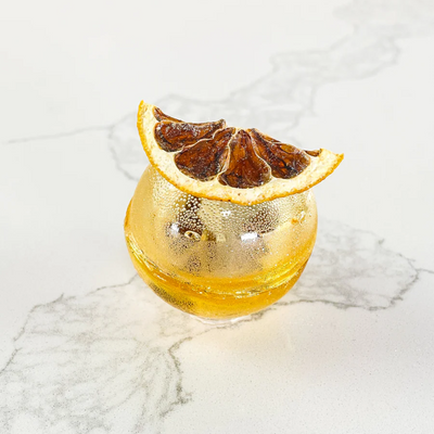 A spherical, orange tea bomb with a dried orange slice on top. The tea bomb is resting on a white marble surface.