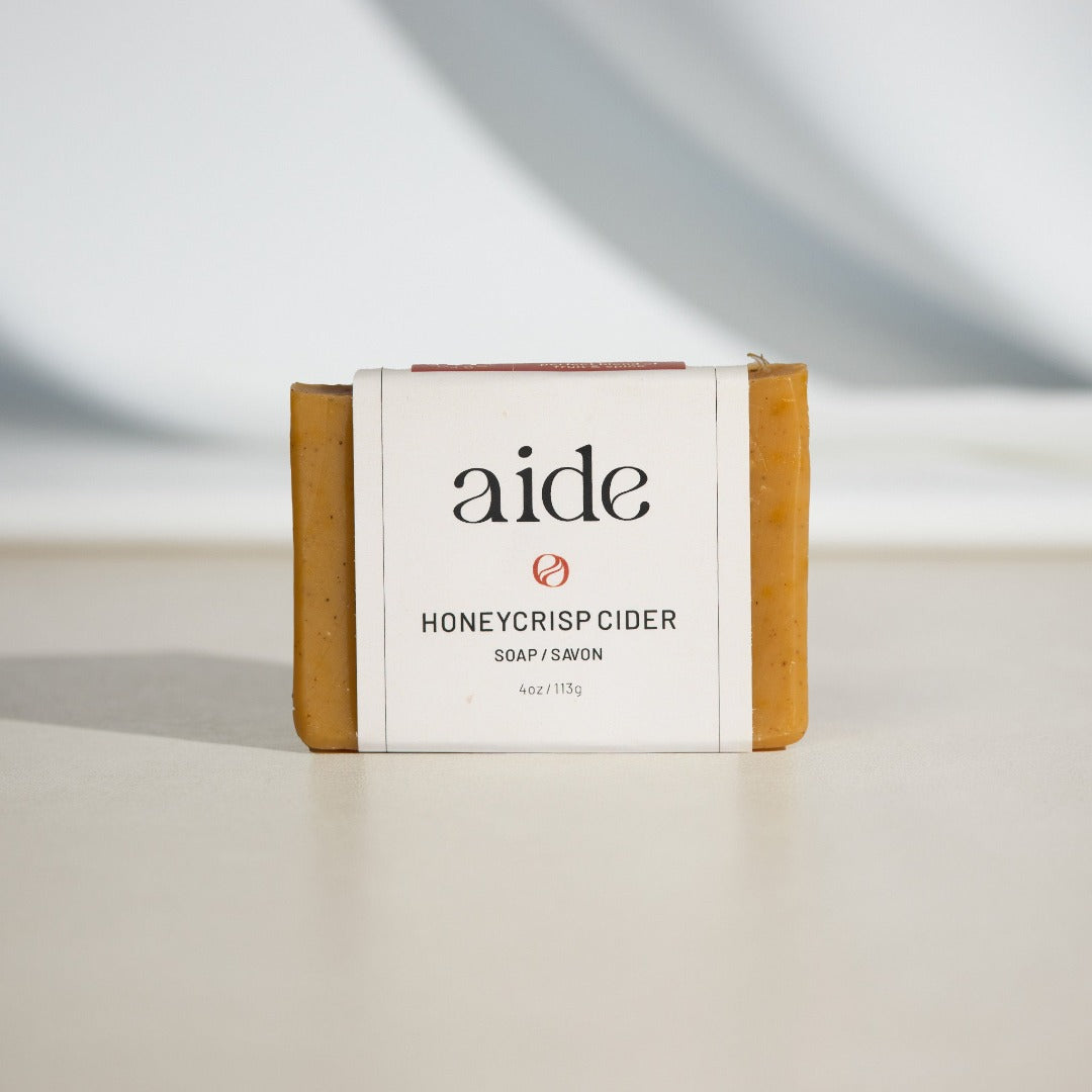 A bar of Aide Bodycare Honeycrisp Cider Soap, featuring a light yellow-orange color and a paper label with the 'aide' logo and 'Honeycrisp Cider' text, displayed on a light-colored surface with a shadow overlay. The soap is natural and handmade, available in Ottawa, Ontario, Canada.