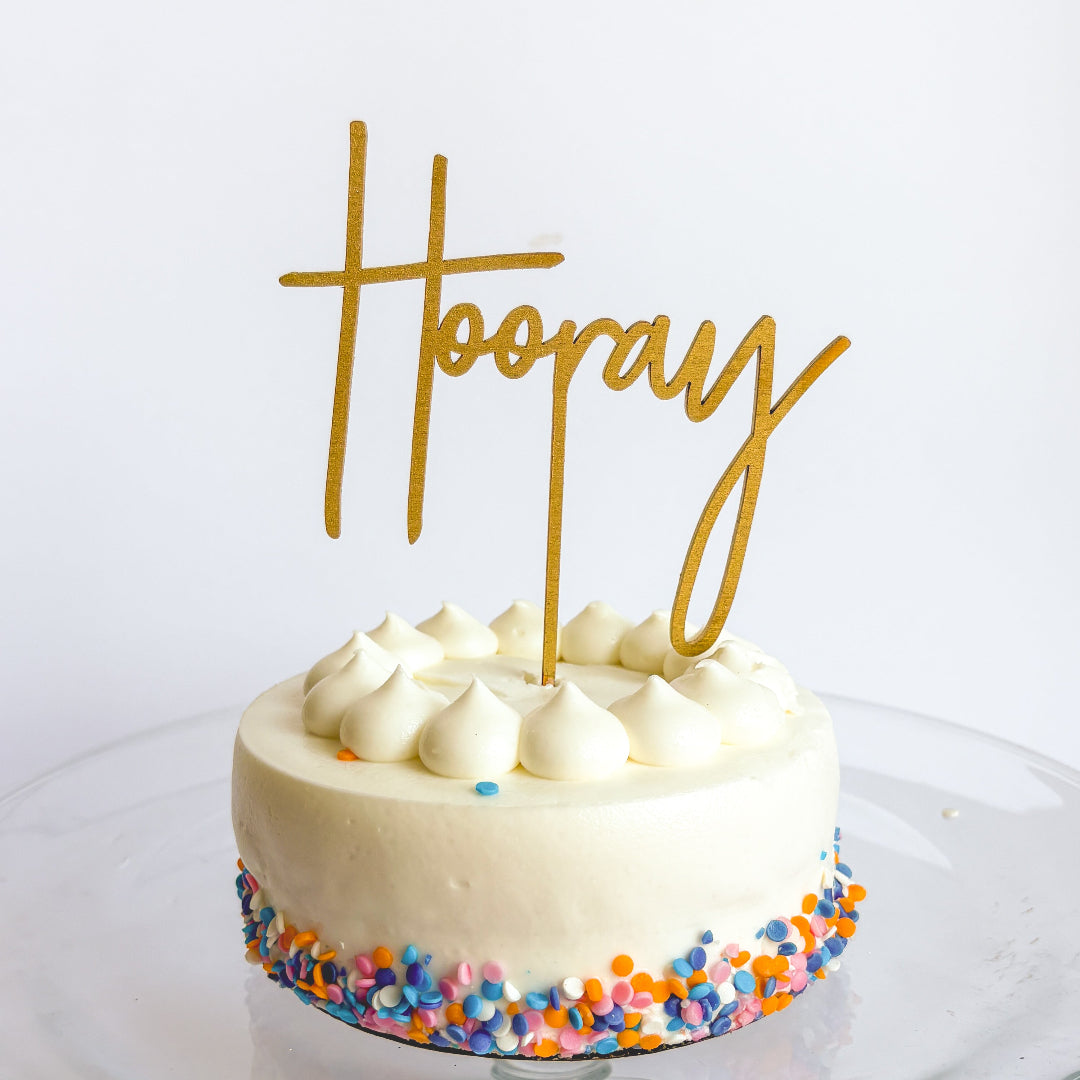 Small round cake with white frosting, colorful sprinkles, and a gold 'Hoopla' topper on a white background.