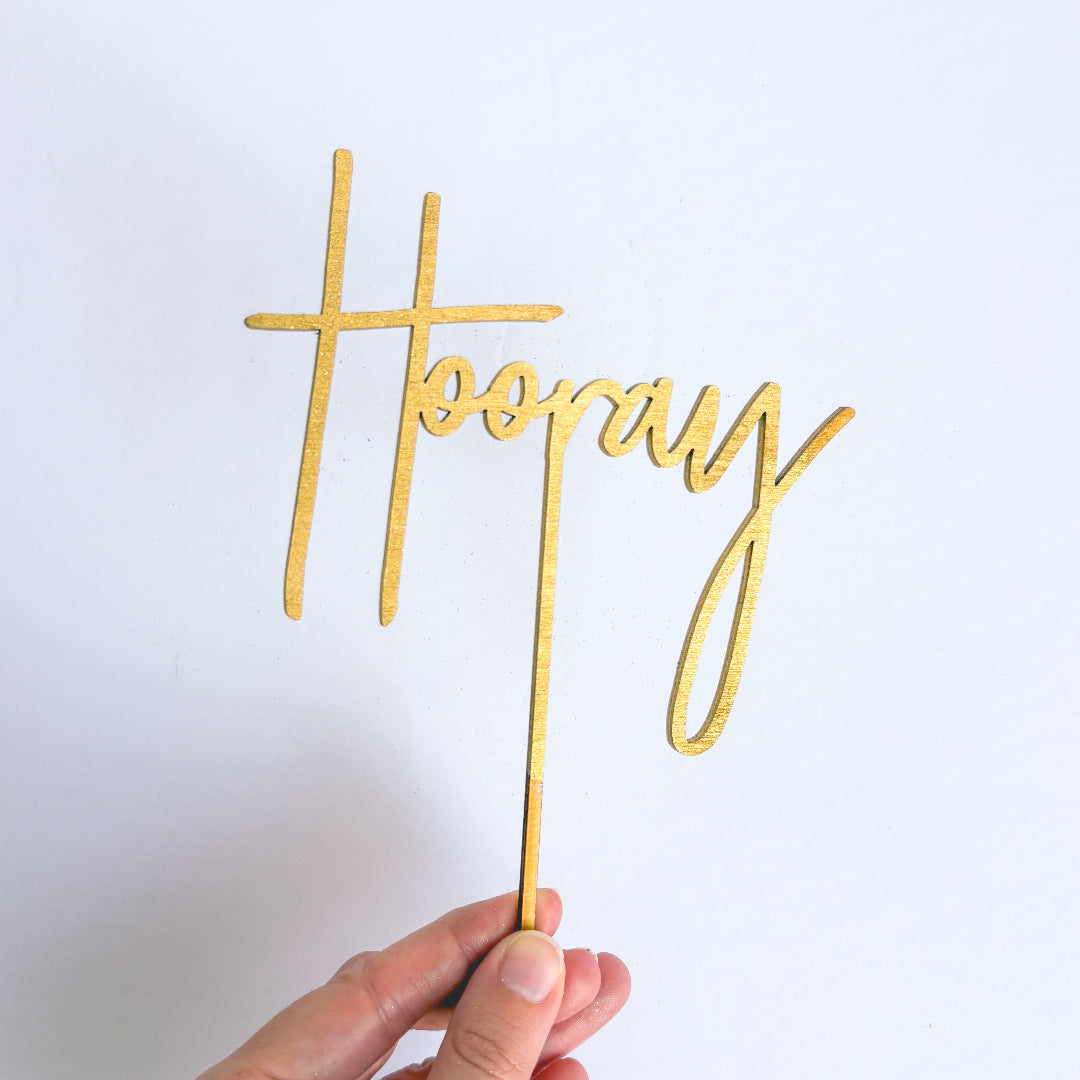 Gold 'Hoopay' cake topper held against a light gray background