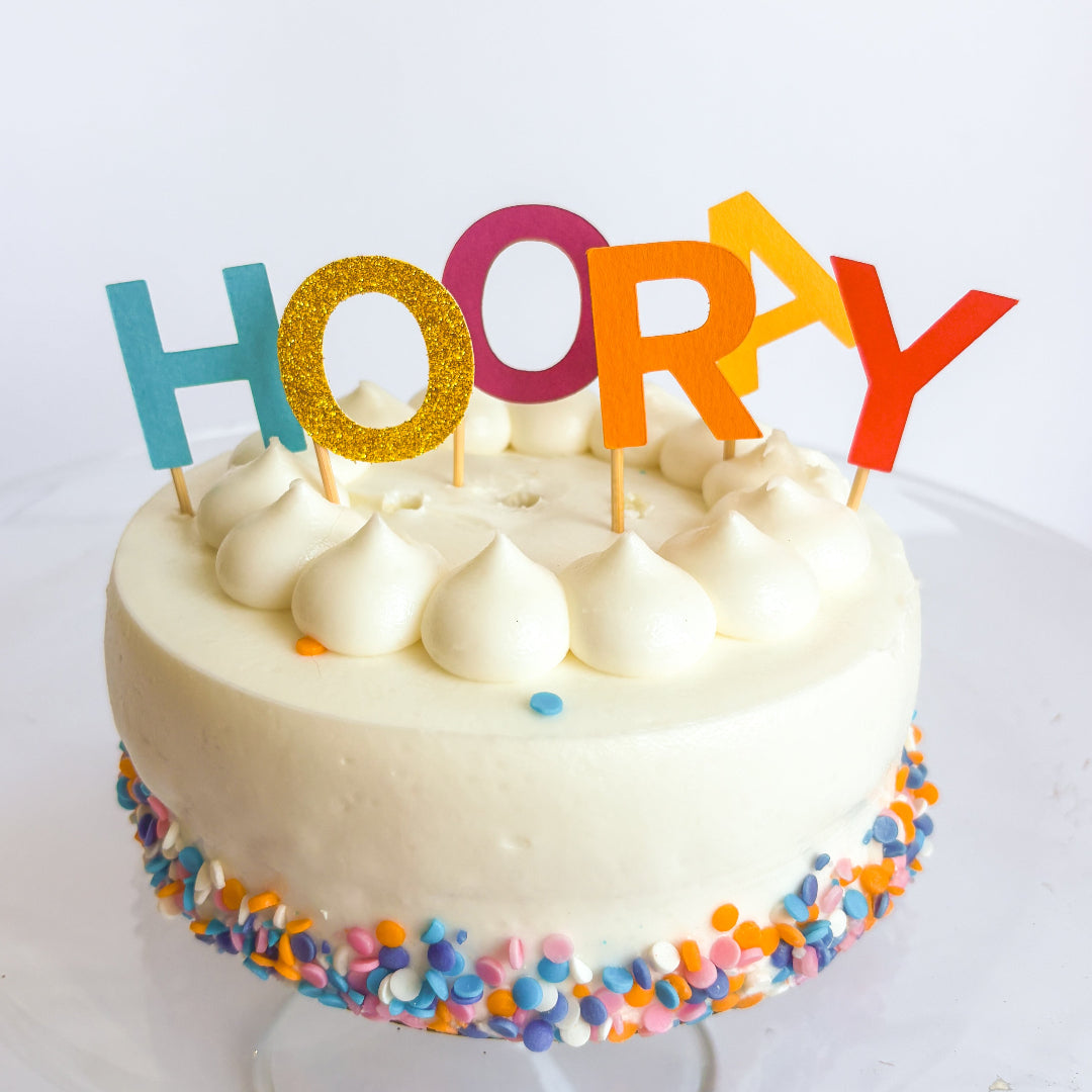 White cake with colorful 'HOORAY' topper on a white background