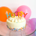 Small white cake with colorful 'HOORAY' topper and balloons in the background