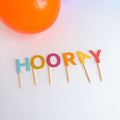 Colorful 'HOORAY' letter candles on toothpicks with a blurred orange object in the background.