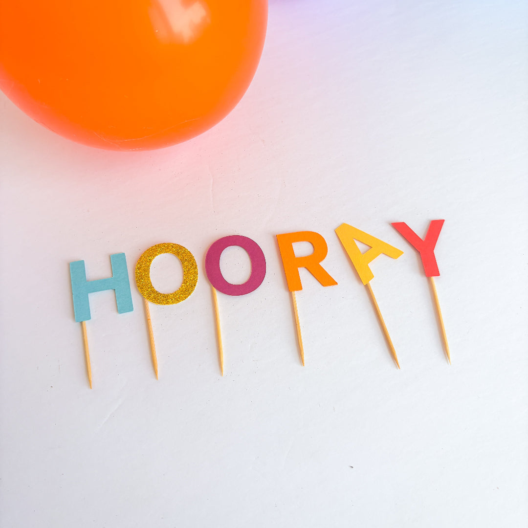 Colorful 'HOORAY' letter candles on toothpicks with a blurred orange object in the background.