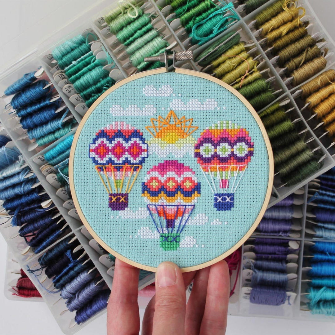 An overhead shot of a finished cross-stitch piece in a wooden embroidery hoop, displayed on a white surface with a potted succulent in the top right corner. The cross-stitch design features three colourful hot air balloons with a sun and clouds against a light blue background.
