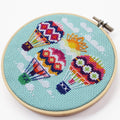 An overhead shot of a finished cross-stitch piece in a wooden embroidery hoop, displayed on a white surface with a potted succulent in the top right corner. The cross-stitch design features three colourful hot air balloons with a sun and clouds against a light blue background.