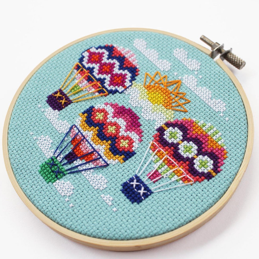 An overhead shot of a finished cross-stitch piece in a wooden embroidery hoop, displayed on a white surface with a potted succulent in the top right corner. The cross-stitch design features three colourful hot air balloons with a sun and clouds against a light blue background.
