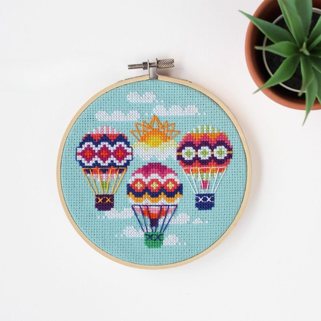An overhead shot of a finished cross-stitch piece in a wooden embroidery hoop, displayed on a white surface with a potted succulent in the top right corner. The cross-stitch design features three colourful hot air balloons with a sun and clouds against a light blue background.