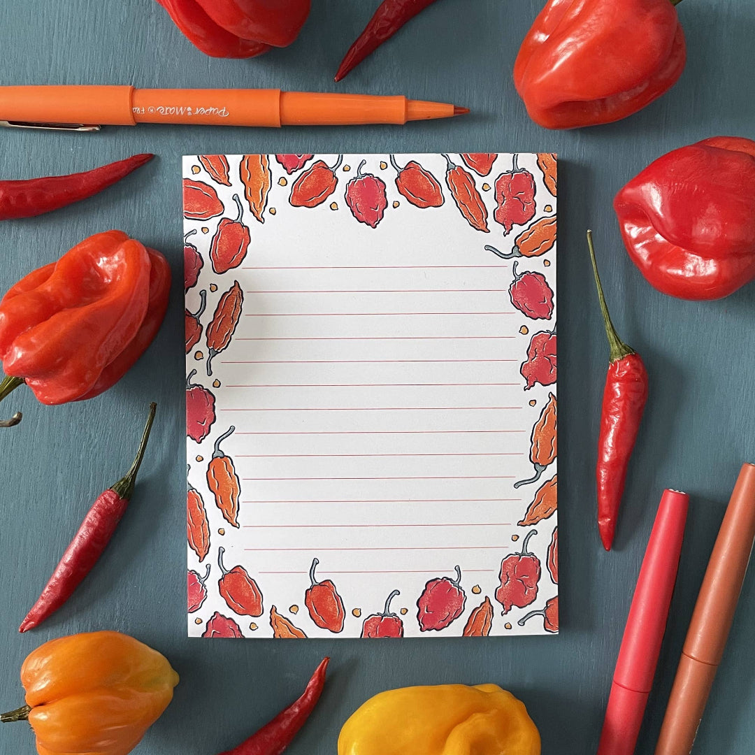 Notepad with chili pepper design surrounded by real red and yellow peppers on a blue background