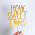 Gold glittery 'HOW SWEET IT IS' cake topper held against a light gray background