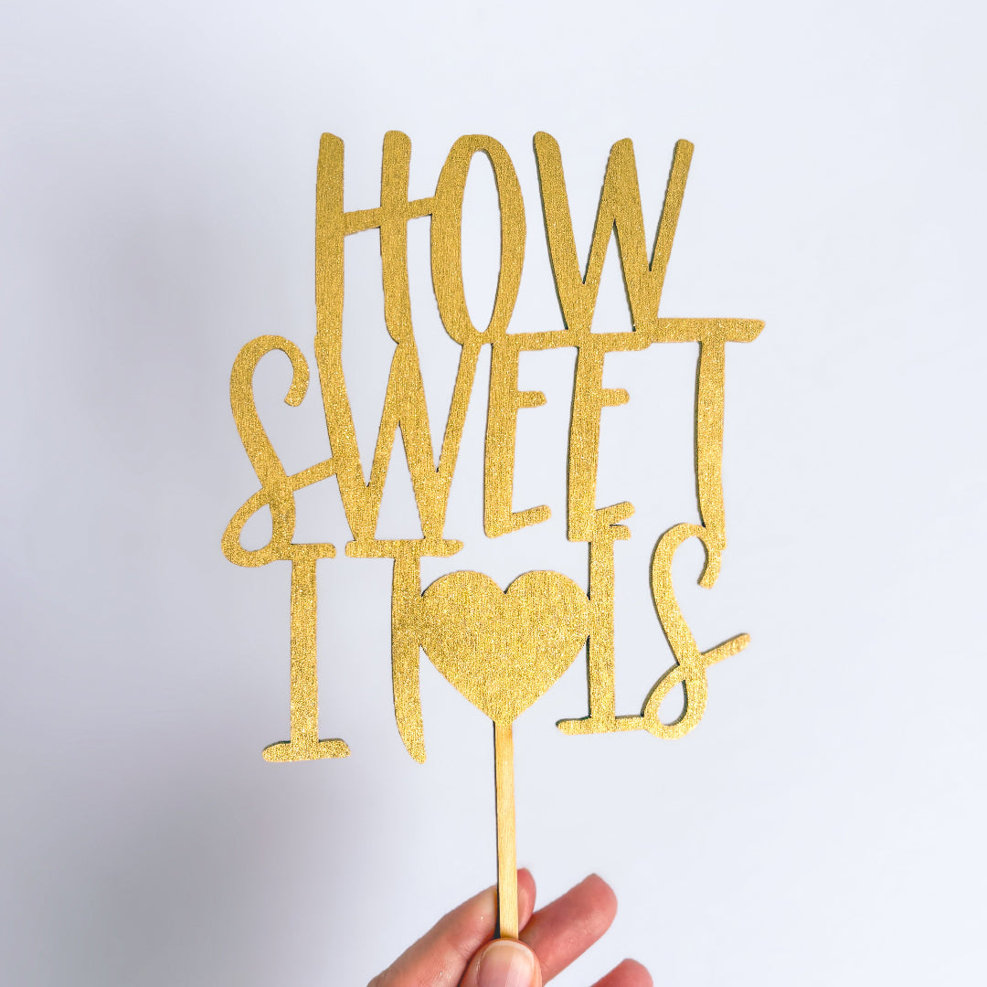 Gold glittery 'HOW SWEET IT IS' cake topper held against a light gray background
