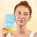 A woman with freckles holds a blue and yellow Cryo facial mask packet next to her face, looking to the side against a light beige background.