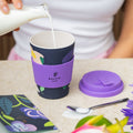 Person pouring milk into a coffee cup with a purple sleeve, surrounded by colorful items on a table.