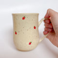 A handmade ceramic mug by Valerie Daoust Ceramics, featuring hand-painted red strawberries on a speckled cream background, displayed on a light gray background. The mug has a large, rounded handle, a speckled texture, and is made in Mississippi Mills, Ontario, Canada.