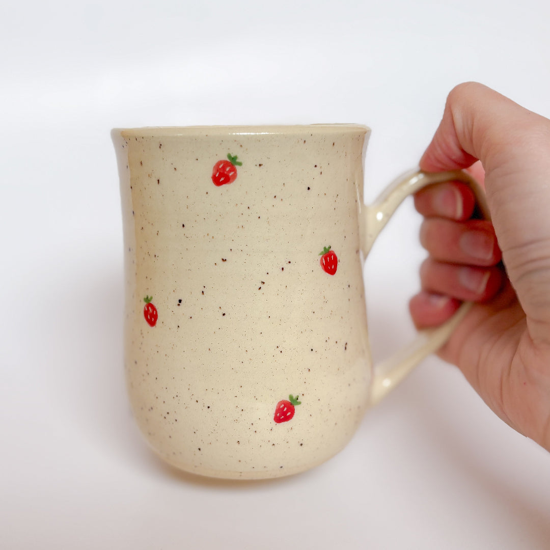 A handmade ceramic mug by Valerie Daoust Ceramics, featuring hand-painted red strawberries on a speckled cream background, displayed on a light gray background. The mug has a large, rounded handle, a speckled texture, and is made in Mississippi Mills, Ontario, Canada.