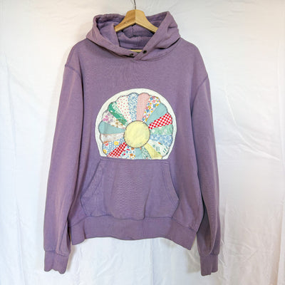 A light purple Bluenotes hoodie, size Large, hanging on a wooden hanger against a white background. The front of the hoodie is visible, with a large, round quilted patchwork sewn onto the chest. The patch has a yellow centre and surrounding fabric strips in a variety of patterns and colours, including light blue, checkered red, and yellow floral prints.