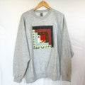 A grey Gildan crewneck sweatshirt, size X-Large, hanging on a wooden hanger against a white background. The front of the sweatshirt is visible, with a square, quilted patchwork panel sewn onto the chest. The patch is made of various fabric scraps in different patterns, including a red and black check and a light blue gingham.
