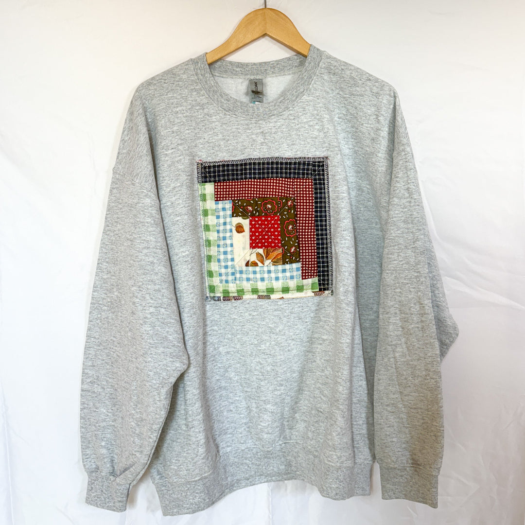 A grey Gildan crewneck sweatshirt, size X-Large, hanging on a wooden hanger against a white background. The front of the sweatshirt is visible, with a square, quilted patchwork panel sewn onto the chest. The patch is made of various fabric scraps in different patterns, including a red and black check and a light blue gingham.