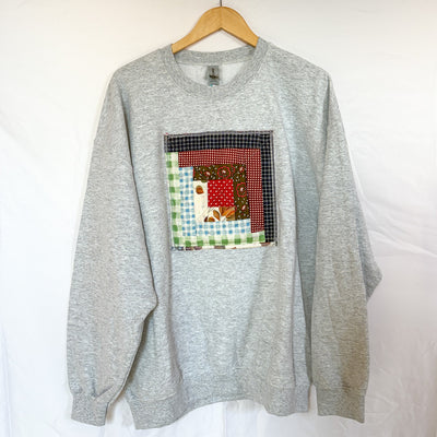A grey Gildan crewneck sweatshirt, size X-Large, hanging on a wooden hanger against a white background. The front of the sweatshirt is visible, with a square, quilted patchwork panel sewn onto the chest. The patch is made of various fabric scraps in different patterns, including a red and black check and a light blue gingham.