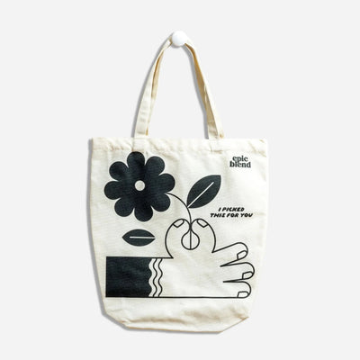 A white tote bag with a black and white illustration. The illustration shows a hand picking a flower. The text 