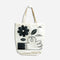 A white tote bag with a black and white illustration. The illustration shows a hand picking a flower. The text 