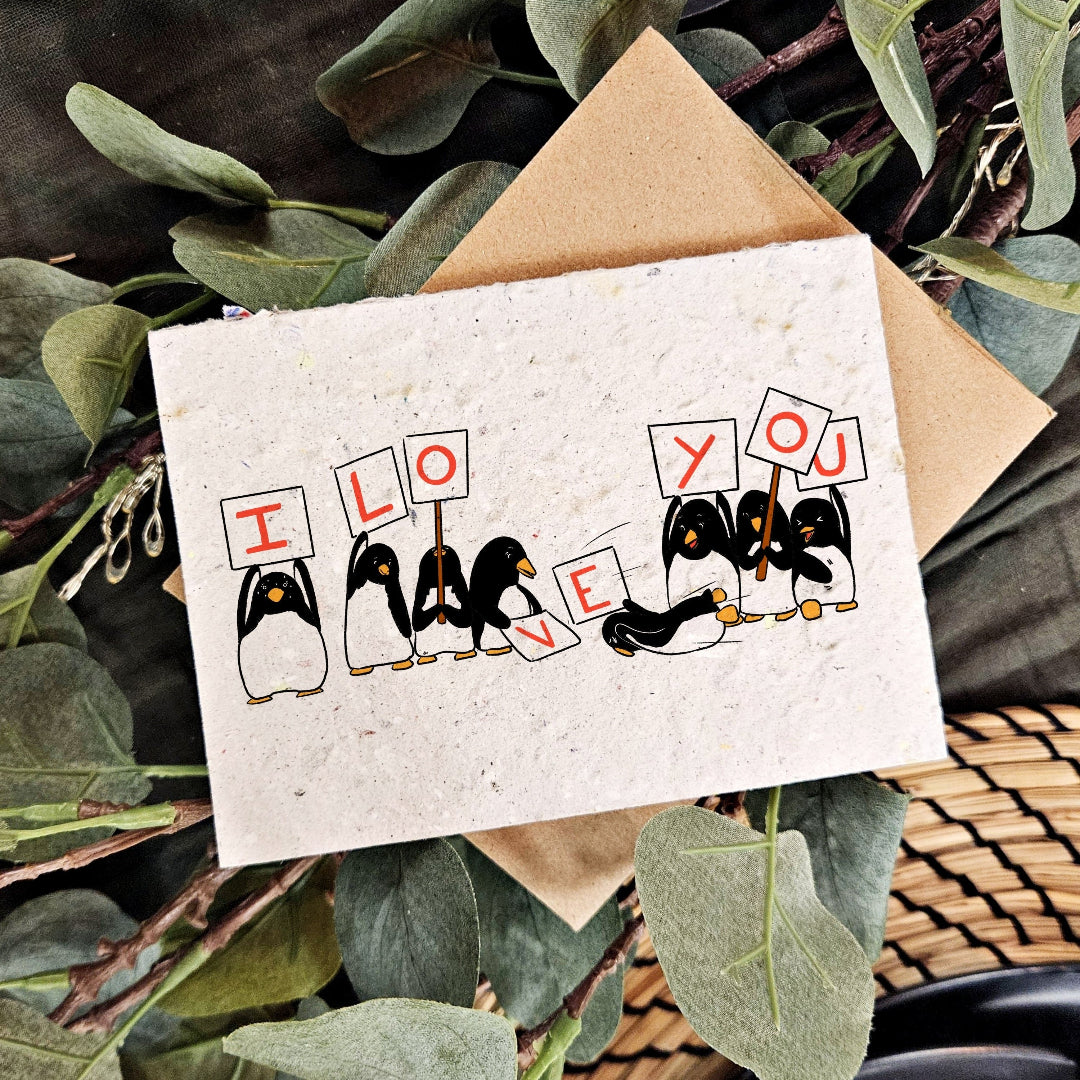 Card with penguins holding signs saying 'I love you' on a natural background.
