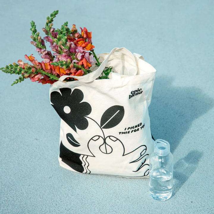 A white tote bag with a black and white illustration. The illustration shows a hand picking a flower. The text 