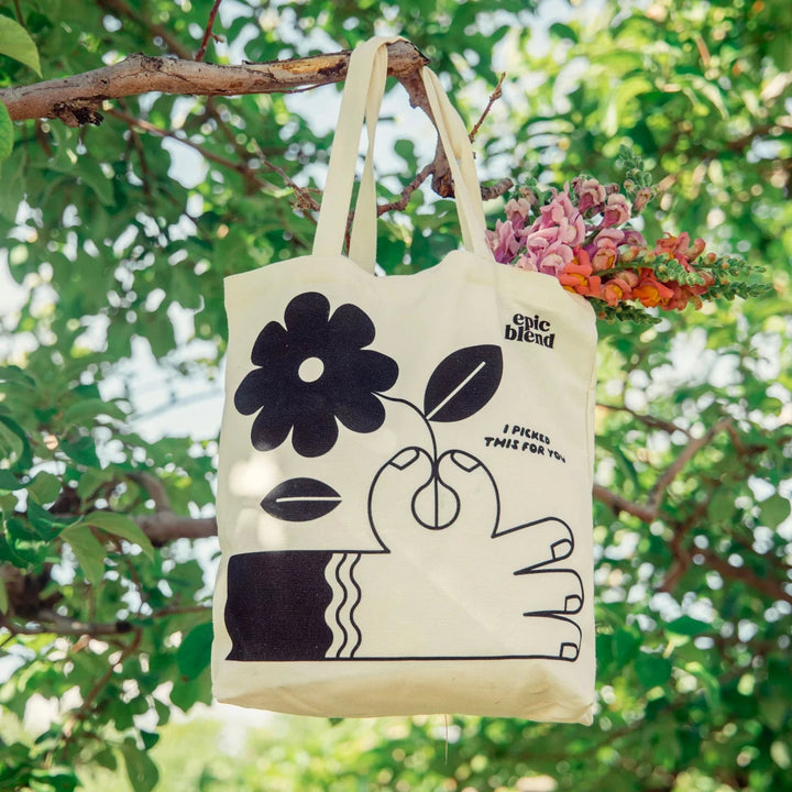 A white tote bag with a black and white illustration. The illustration shows a hand picking a flower. The text 