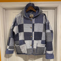 A hooded patchwork quilted jacket in navy blue, white, and gray striped fabrics, hanging on a wooden hanger against a white paneled door.
