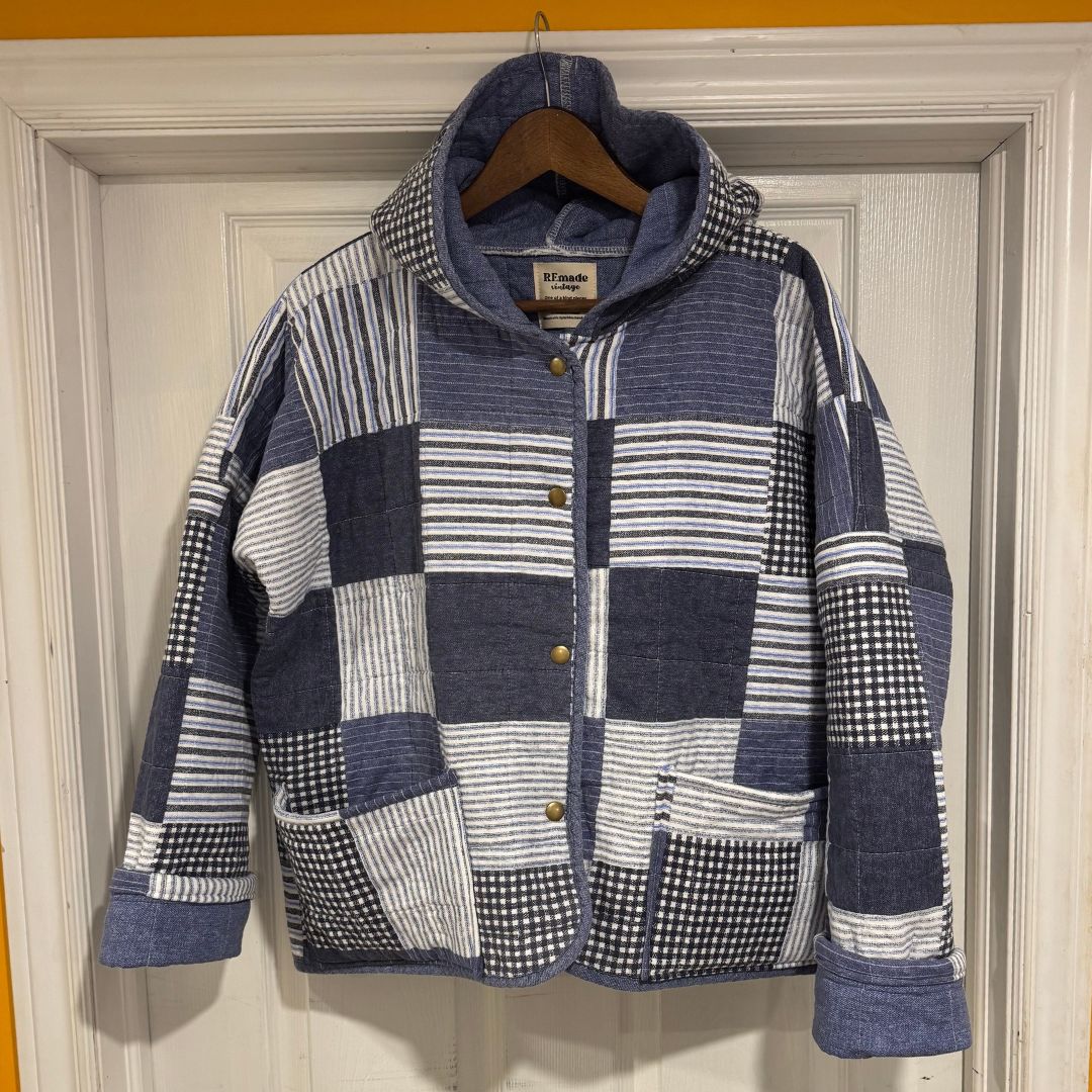 A hooded patchwork quilted jacket in navy blue, white, and gray striped fabrics, hanging on a wooden hanger against a white paneled door.