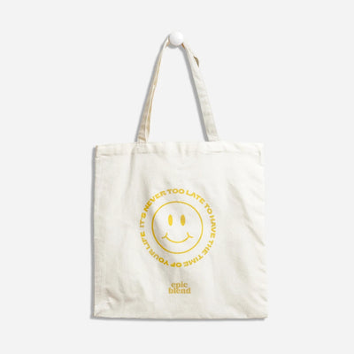 A white tote bag with a yellow smiley face design. The text around the smiley face reads: 