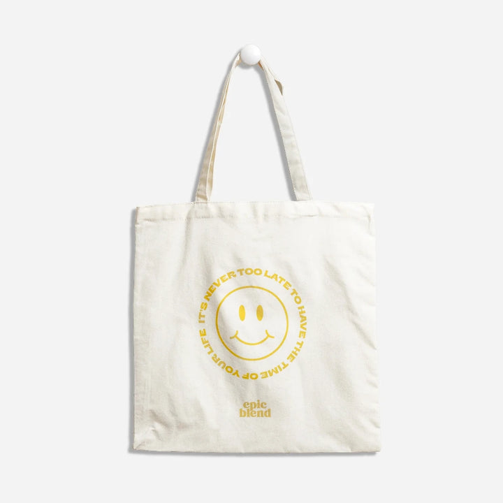 A white tote bag with a yellow smiley face design. The text around the smiley face reads: 