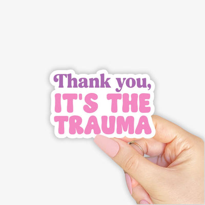 Sticker with 'Thank you, it's the trauma' text held by a hand on a light pink background