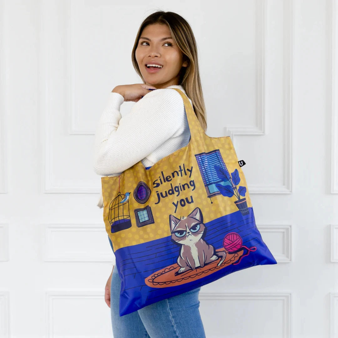 A blue and yellow tote bag with a humorous illustration of a grumpy cat sitting on a rug and silently judging the viewer. The text "Silently Judging You" is printed on the bag.