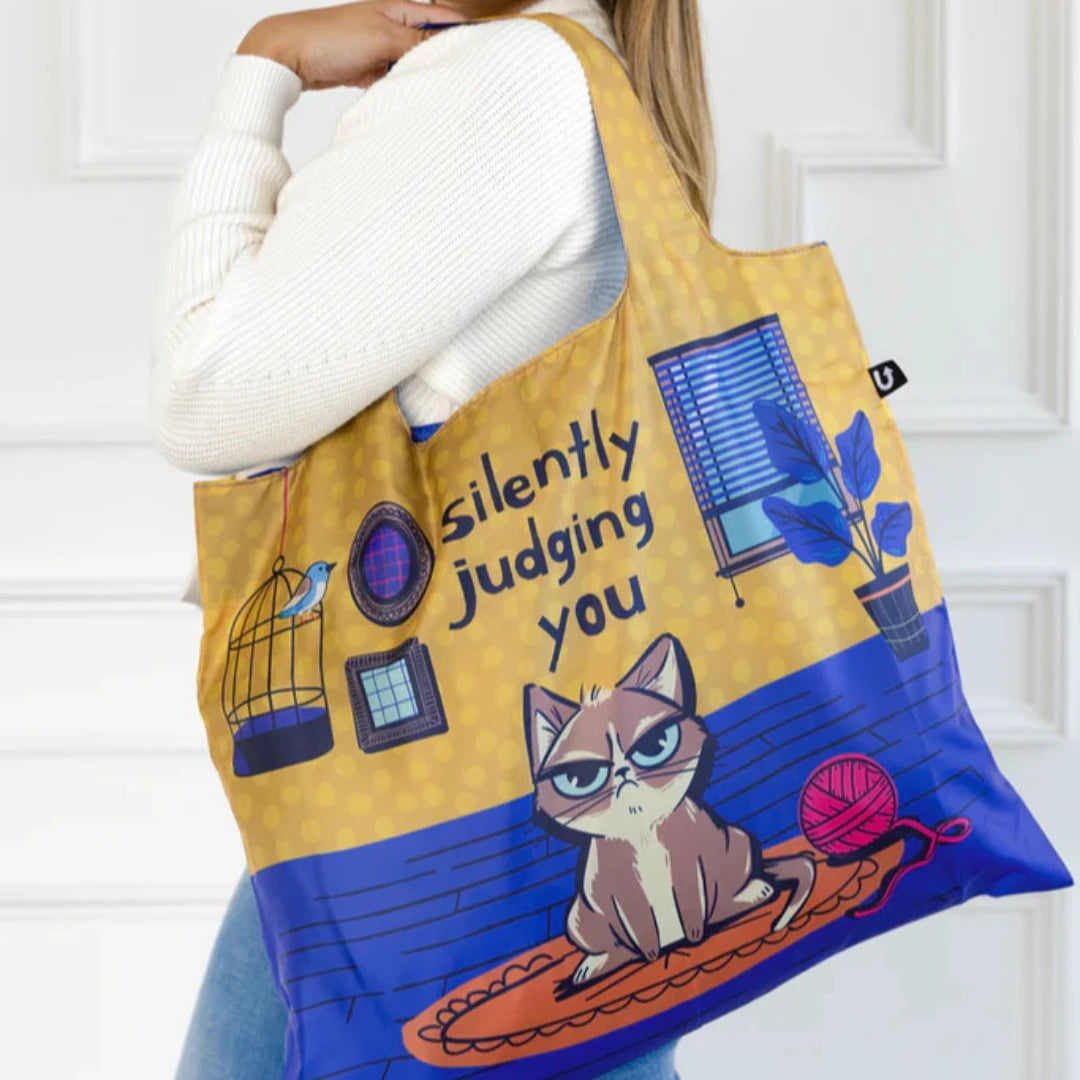 A blue and yellow tote bag with a humorous illustration of a grumpy cat sitting on a rug and silently judging the viewer. The text "Silently Judging You" is printed on the bag.