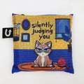 A blue and yellow tote bag with a humorous illustration of a grumpy cat sitting on a rug and silently judging the viewer. The text "Silently Judging You" is printed on the bag.