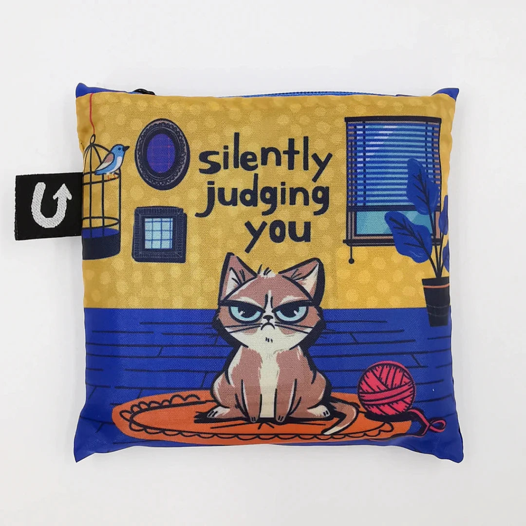 A blue and yellow tote bag with a humorous illustration of a grumpy cat sitting on a rug and silently judging the viewer. The text "Silently Judging You" is printed on the bag.