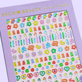 A sheet of colorful nail art stickers with various shapes like rainbows, mushrooms, flowers, and hearts. The sheet is purple and has the brand name "Deco Beauty" printed on it. There's a section titled "JELLY" and instructions on how to apply the stickers.
