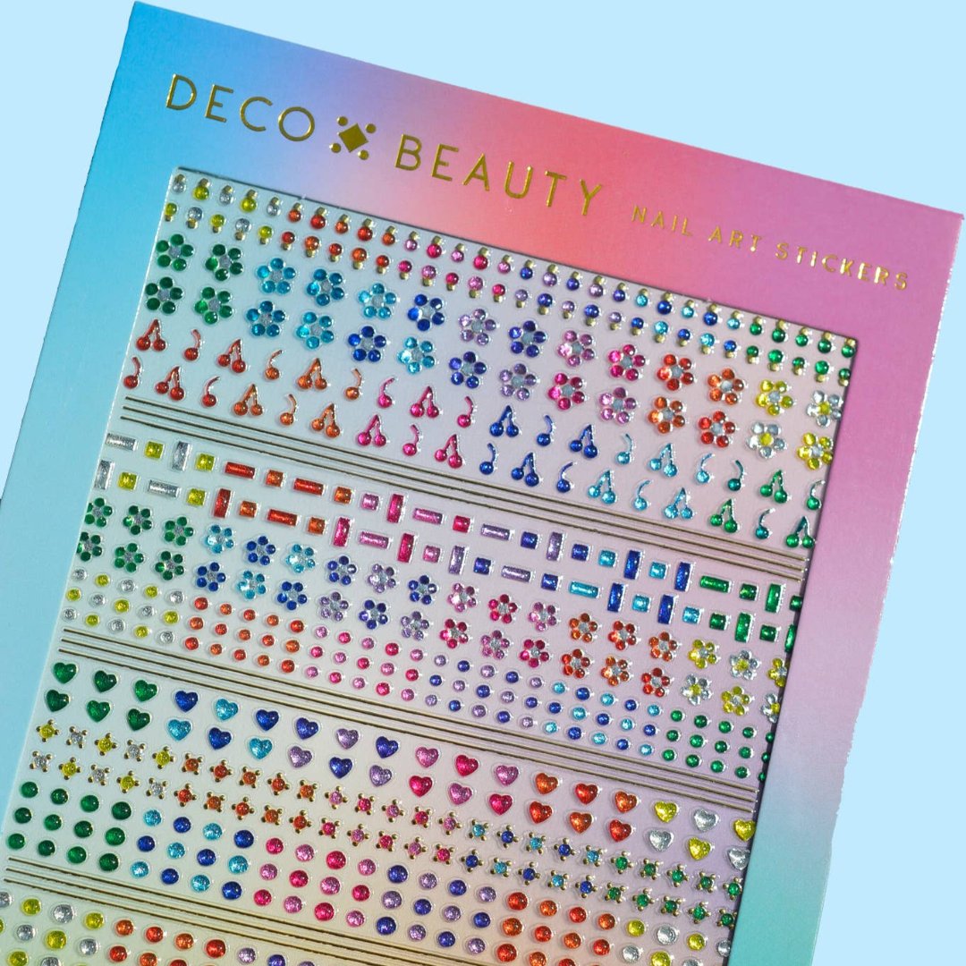 A sheet of colorful nail art stickers with various shapes like stars, hearts, flowers, and geometric designs. The sheet is holographic and has the brand name "Deco Beauty" printed on it. There's a section titled "JEWELS" and instructions on how to apply the stickers.