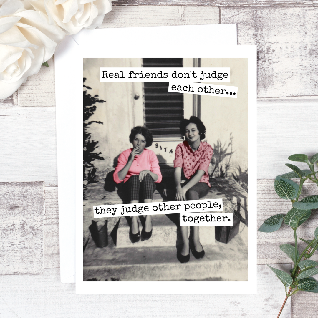 A greeting card featuring a vintage black and white photo of two women sitting on steps, one smoking. Text on the card reads: "Real friends don't judge each other... they judge other people, together."
