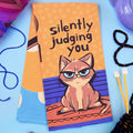 Towel with a cat illustration and text 'silently judging you' on an orange background.