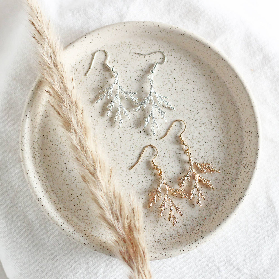 Gold leaf earrings on a speckled ceramic plate with a dried plant stem.