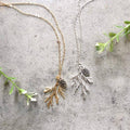 Two necklaces on a grey surface. One necklace has a gold-colored pine cone and branch pendant, and the other has a silver-colored pine cone and branch pendant. Both necklaces have delicate chains.