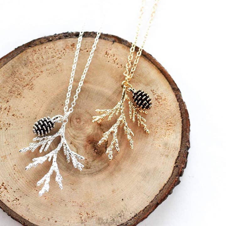 Two necklaces on a grey surface. One necklace has a gold-colored pine cone and branch pendant, and the other has a silver-colored pine cone and branch pendant. Both necklaces have delicate chains.